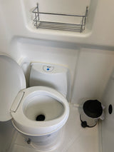 This compact RV rental bathroom features a white toilet, an overhead wire shelf, and a small waste bin on the floor.