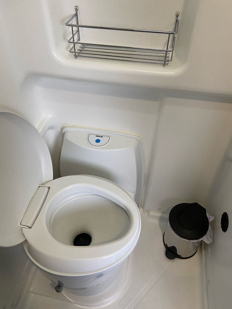 This compact RV rental bathroom features a white toilet, an overhead wire shelf, and a small waste bin on the floor.