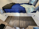 Cozy RV rental interior features a bed with blue comforter & patterned blanket, plus dark grey seating cushions.