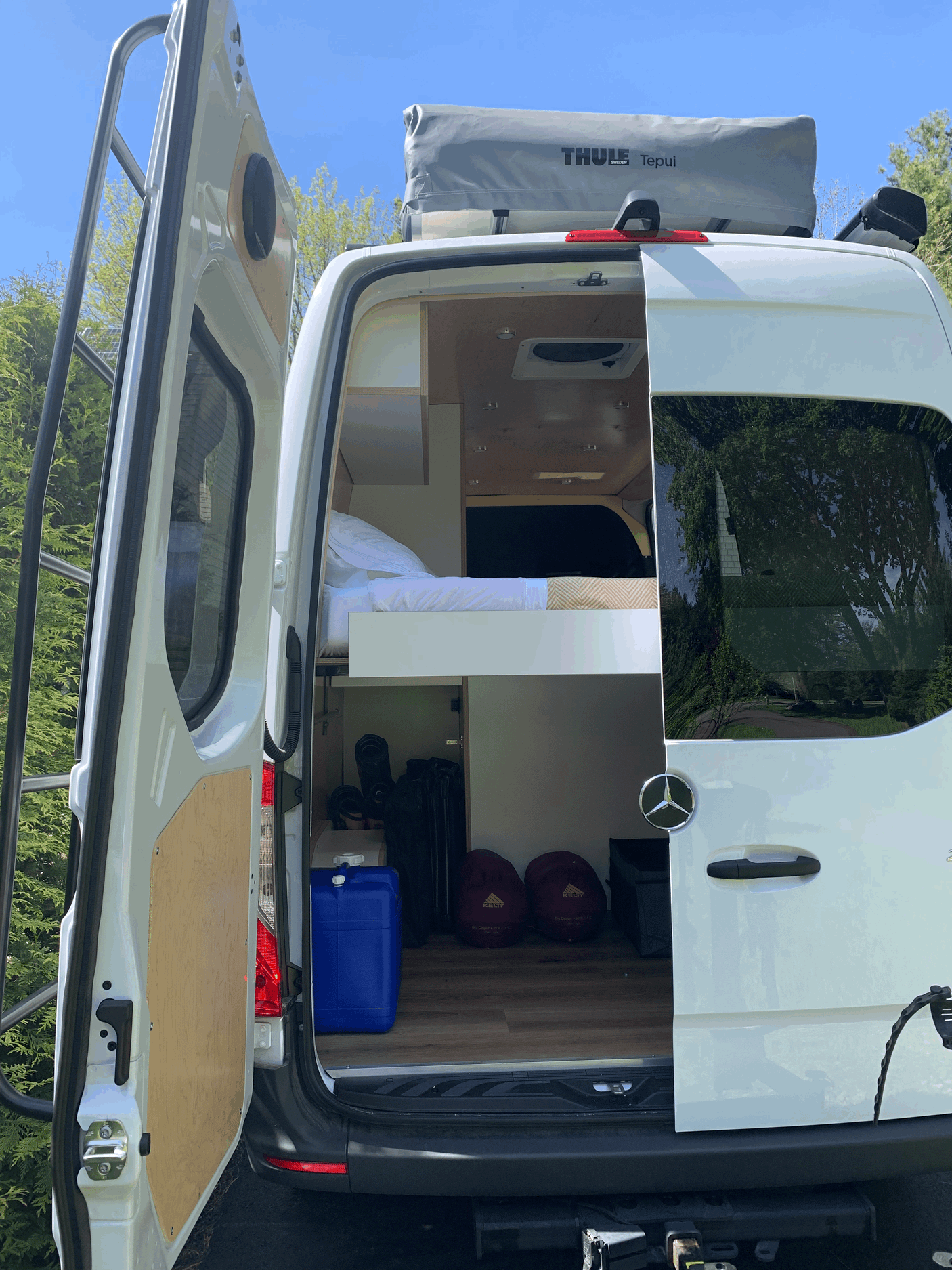Open doors of a white Mercedes Sprinter camper van, ideal for RV rental. Shows elevated bed, storage area, and rooftop tent.