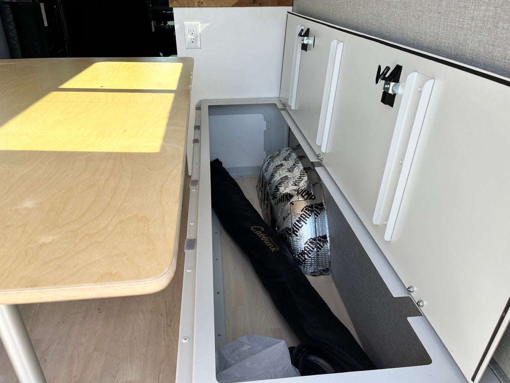 RV rental interior: practical under-bench storage with Kilmat insulation, a black bag, and clean white cabinets.