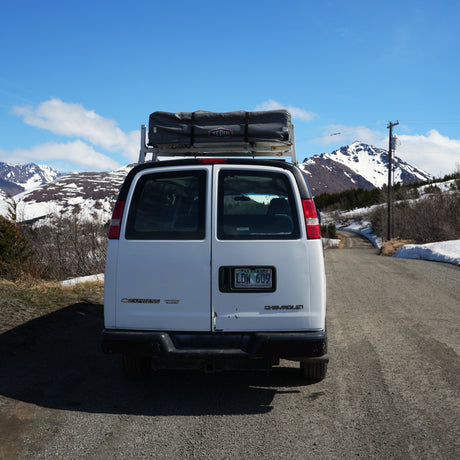 White cargo van RV rental with rooftop tent on a scenic gravel road in snowy Alaskan mountains under blue sky.