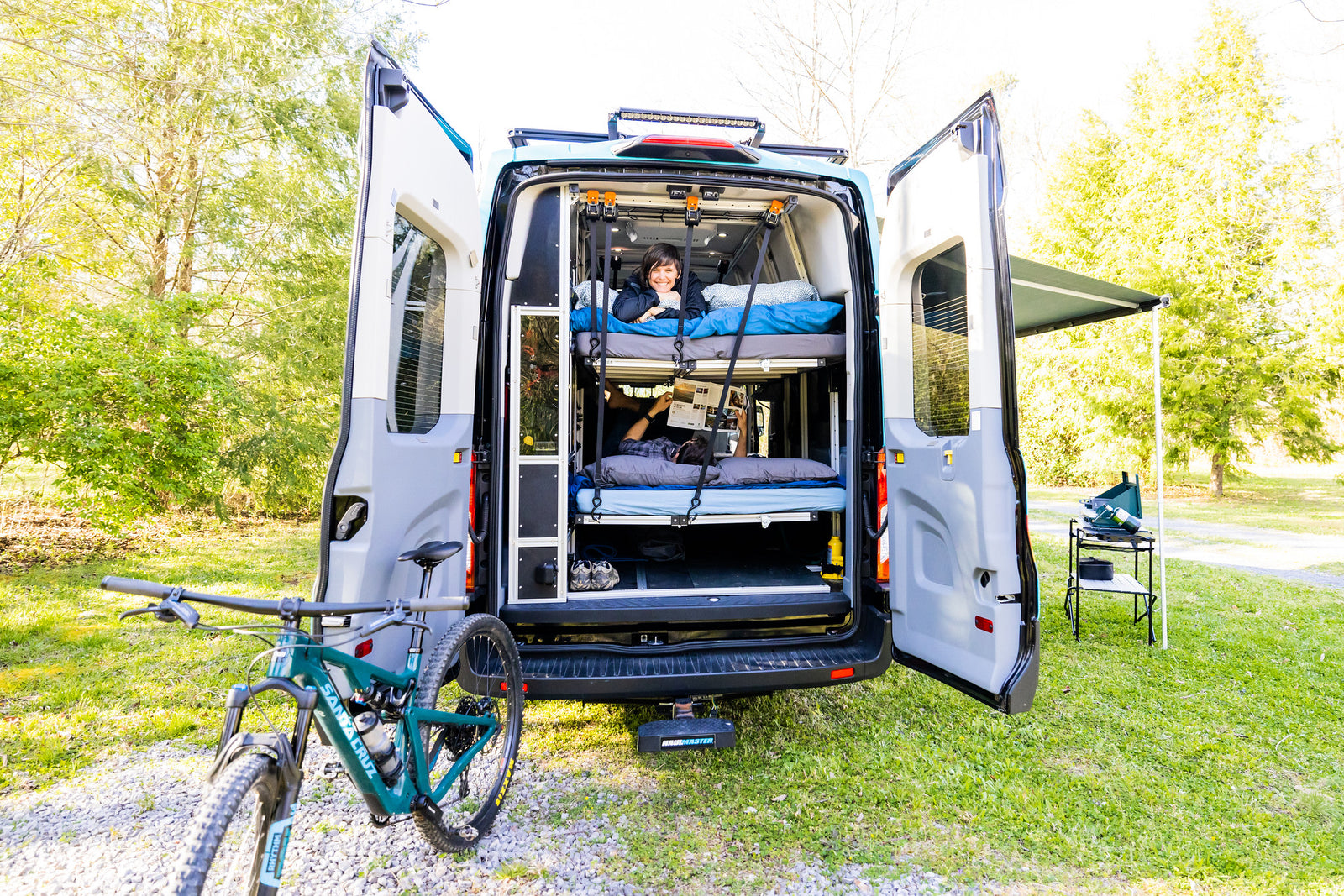 Open RV rental camper van reveals bunk beds with people relaxing, plus a mountain bike & outdoor cooking setup.