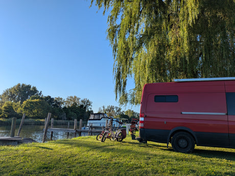 Scenic rv rental setup: Red camper van, bikes, and boat "Lady May" by a tranquil waterway and green trees.