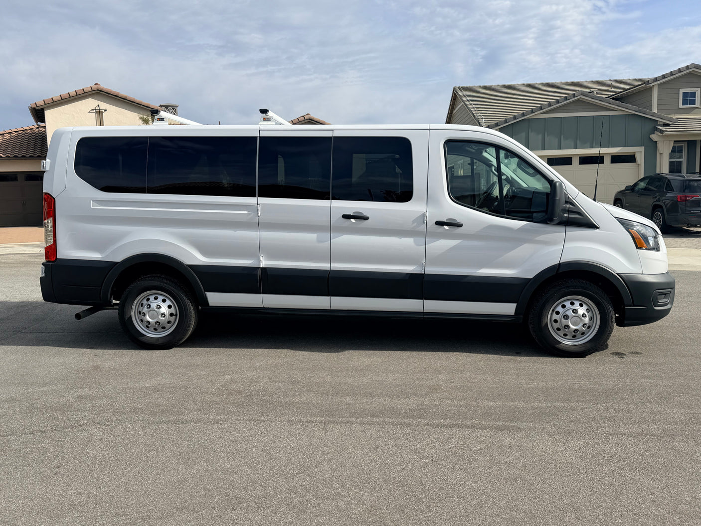 White Ford Transit passenger van, a potential RV rental or conversion, parked on a residential street under a cloudy sky.