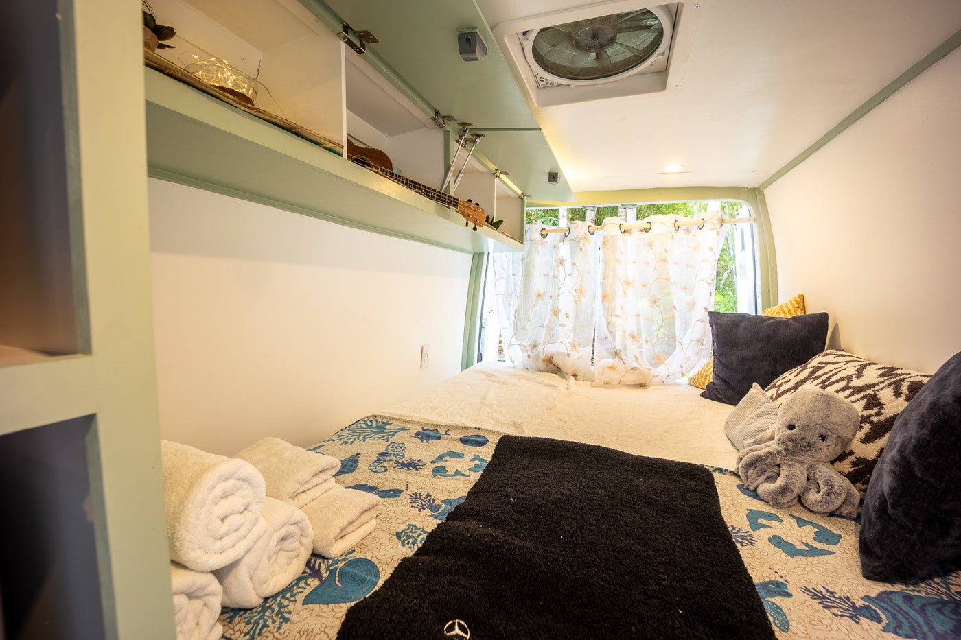 Cozy RV rental interior: bed, overhead storage with a ukulele, window with curtains, and ceiling fan. — photo 4