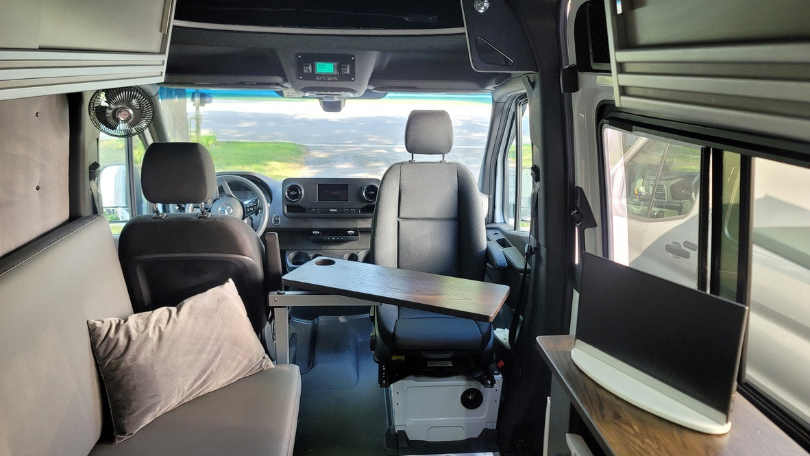 View of a modern RV rental interior featuring swivel seats, a portable table, overhead storage, and a comfy bench.