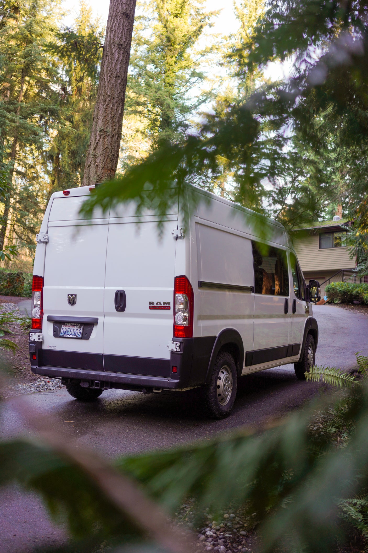 Rear view of a white Ram ProMaster van, an rv rental, on a shaded forest driveway, framed by foliage.