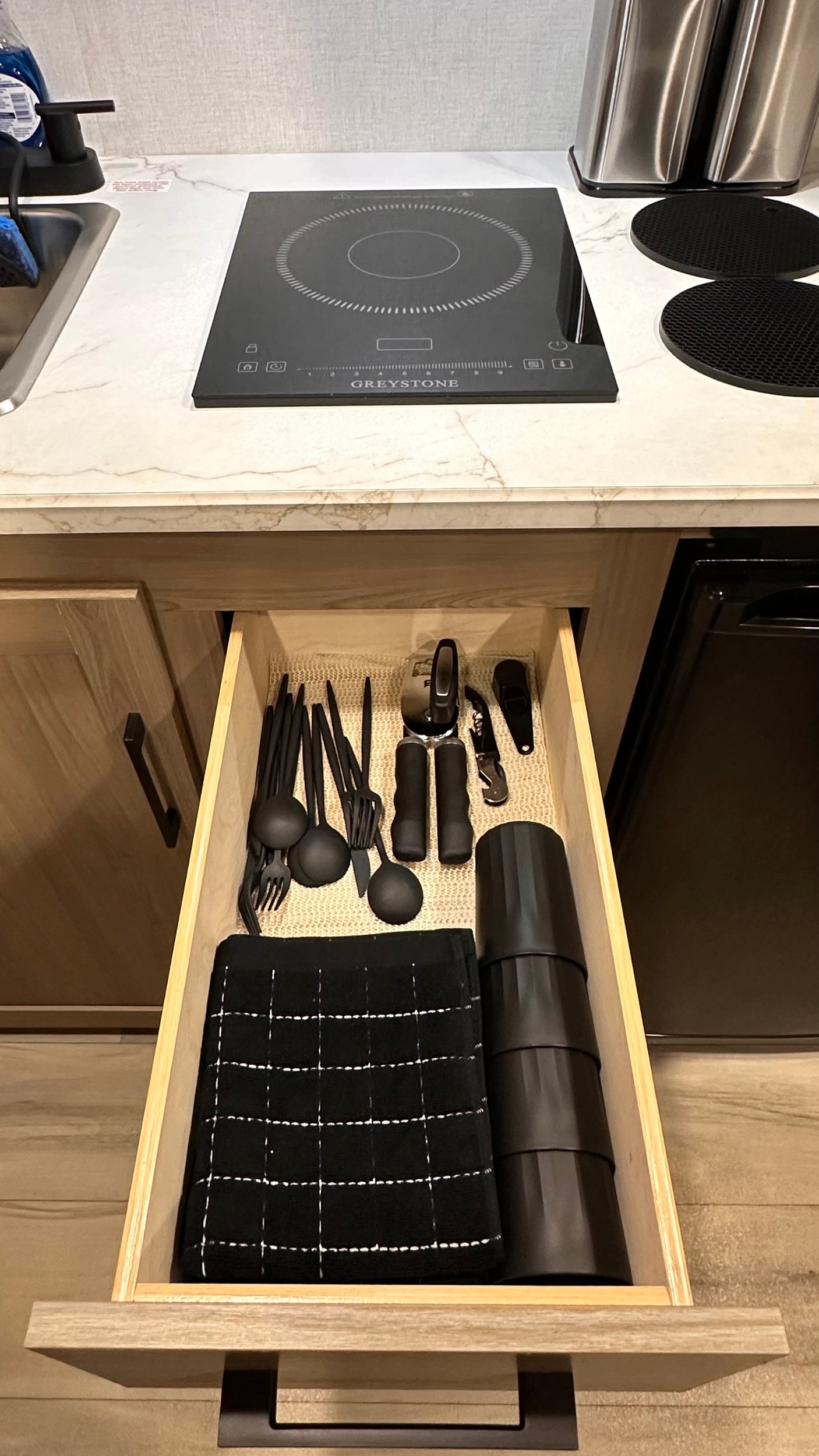RV rental kitchen: Induction cooktop, sink, and a well-organized drawer with black utensils, cups, and a towel.