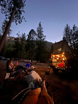 Twilight camping scene: People relaxing at an RV rental site. Rooftop tent, vehicle, and festive lights illuminating the forest campsite.