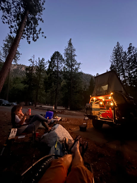 Twilight camping scene: People relaxing at an RV rental site. Rooftop tent, vehicle, and festive lights illuminating the forest campsite.
