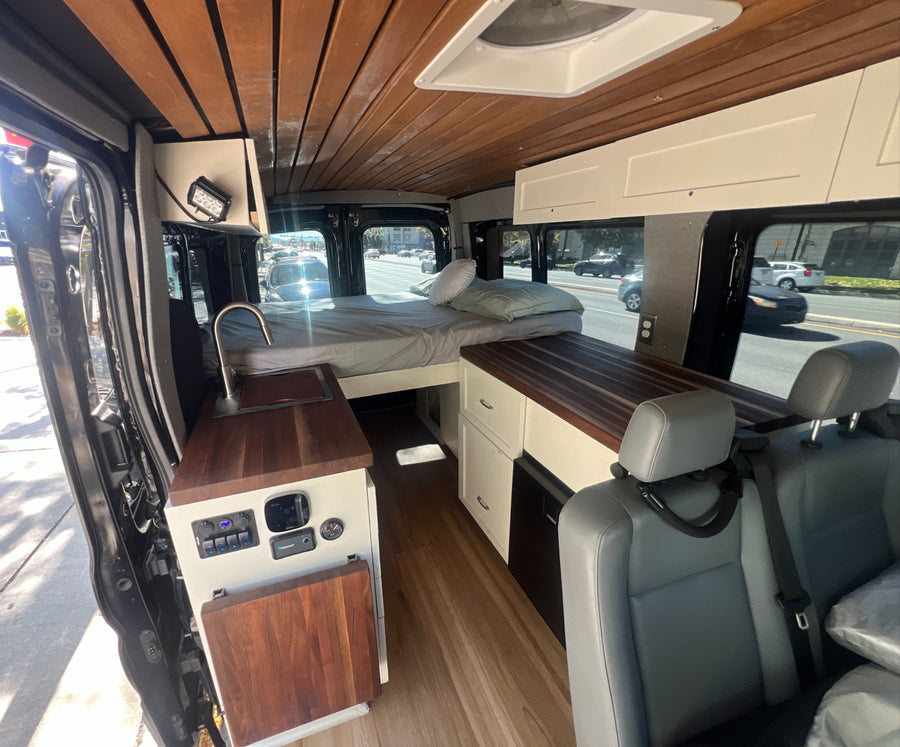 Interior of a modern rv rental camper van, featuring a cozy bed, kitchenette, wood ceiling, and ample storage.