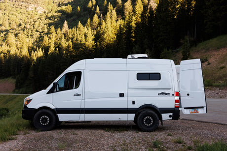 White **rv rental** camper van with open rear doors, parked on a dirt pull-off against a backdrop of sunlit forested mountains.