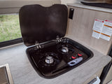 RV rental kitchen showing an open two-burner gas stove, a lighter, a sink, and essential safety labels on the wall.