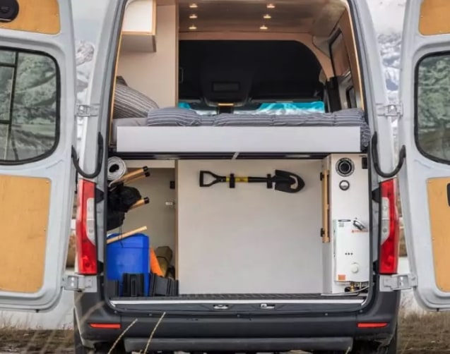 parked on a vast salt flat. Ideal for remote travel adventures.|This rv rental shows a well-organized camper van interior with a bed above storage for gear like a shovel & water heater — photo 2