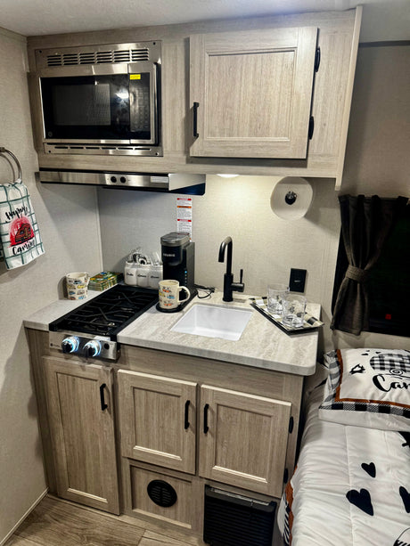Bright RV rental kitchen: microwave, two-burner stove, sink, coffee maker. Cozy bed and decor nearby for a comfy stay.
