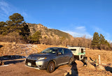 A grey SUV tows a Sunray RV rental through a mountainous, dry landscape with sparse snow patches under blue sky.