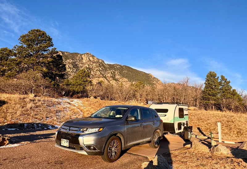 A grey SUV tows a Sunray RV rental through a mountainous, dry landscape with sparse snow patches under blue sky. — photo 7