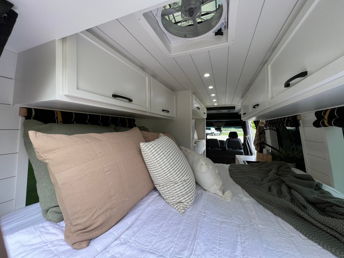 Cozy camper van interior of an RV rental, featuring a large bed with pillows, sleek white cabinets, and overhead lighting. — photo 15