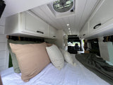 Cozy camper van interior of an RV rental, featuring a large bed with pillows, sleek white cabinets, and overhead lighting.