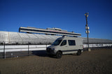 Geotrek RV rental adventure van at New Hampshire Motor Speedway, featuring empty grandstands under a clear blue sky.