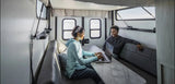 Spacious RV rental interior: two women, one on laptop at table, other relaxing, with scenic outdoor views from windows.