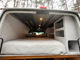 Inside an rv rental van conversion: a cozy bed under a stylish wood plank ceiling with spotlights & built-in storage units.