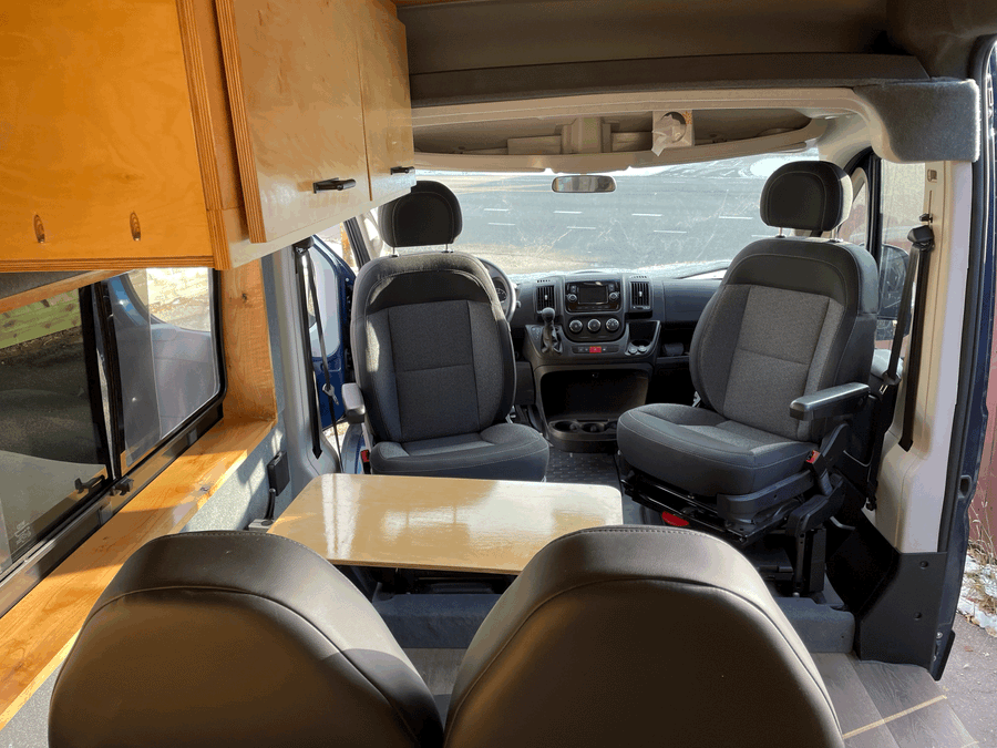 Inside this **rv rental** camper van: front swivel seats, a dining table, and wooden overhead storage for your journey.
