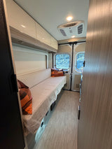 Bright rv rental camper van interior: long bench seat, overhead storage, and rear windows revealing an outdoor scene.