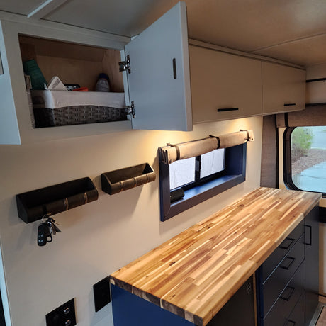 This RV rental features a modern interior with light cabinets, open storage, a window with shade, and a butcher block counter.