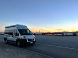 Silver RV rental camper van parked at sunset beside a long train & wind turbines.