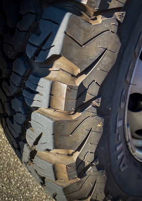Close-up of a rugged all-terrain tire with visible new mold 'hairs,' ready for an RV rental journey. Strong sunlight highlights the tread.