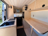 Spacious rv rental interior featuring a wood-paneled van conversion with a kitchenette, bed, and work counter for comfortable travel.