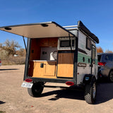 This small RV rental camper has its rear hatch open, revealing a kitchenette with a microwave, sink, and fridge, parked outdoors.