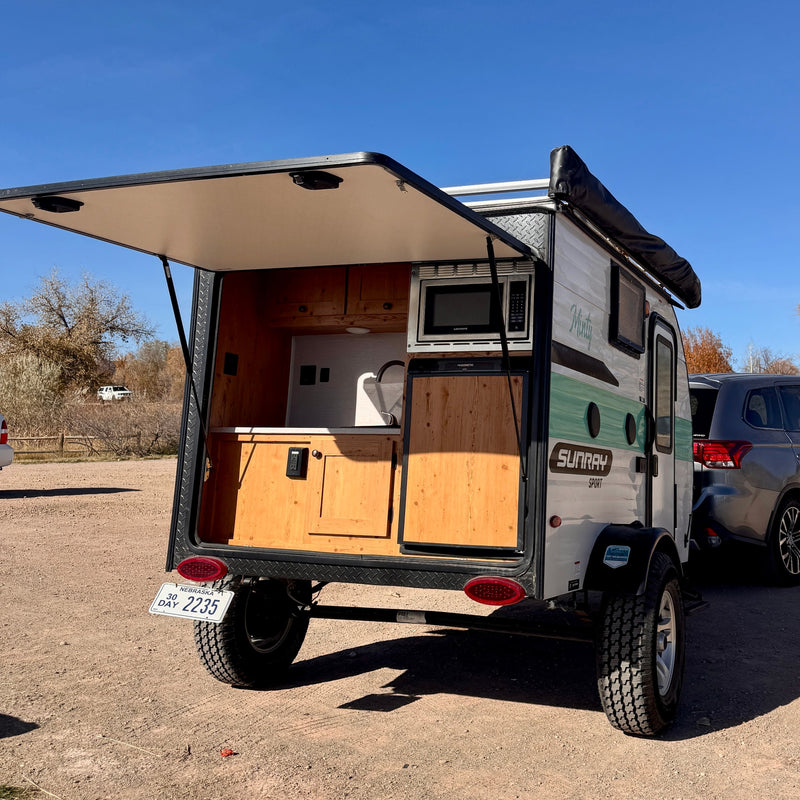 This small RV rental camper has its rear hatch open, revealing a kitchenette with a microwave, sink, and fridge, parked outdoors. — photo 6