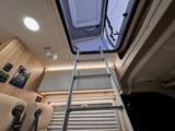 RV rental interior. A ladder ascends to an open overhead bunk/loft, with a control panel and warm lighting visible.