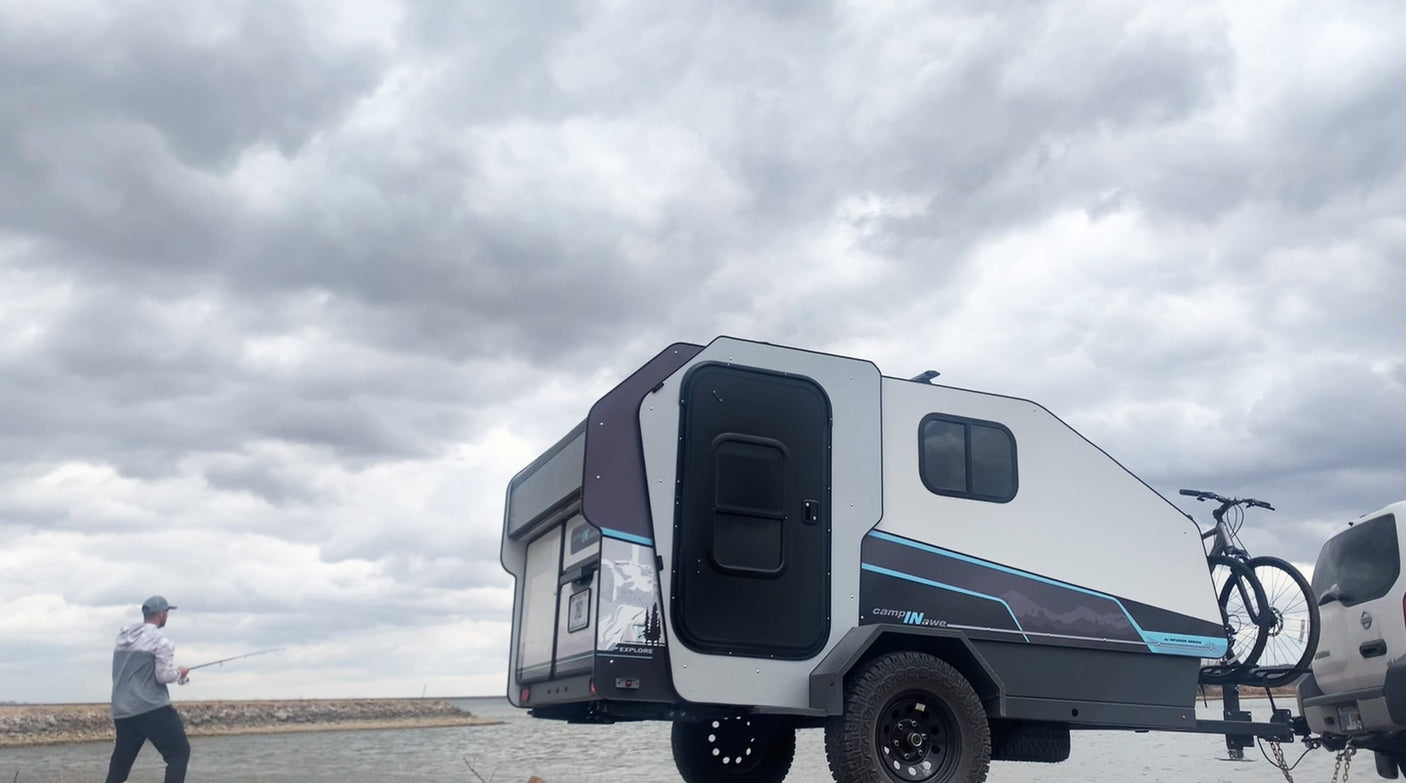 Explore with an RV rental! A man fishes by water, while a modern compact camping trailer with a bike is parked nearby.