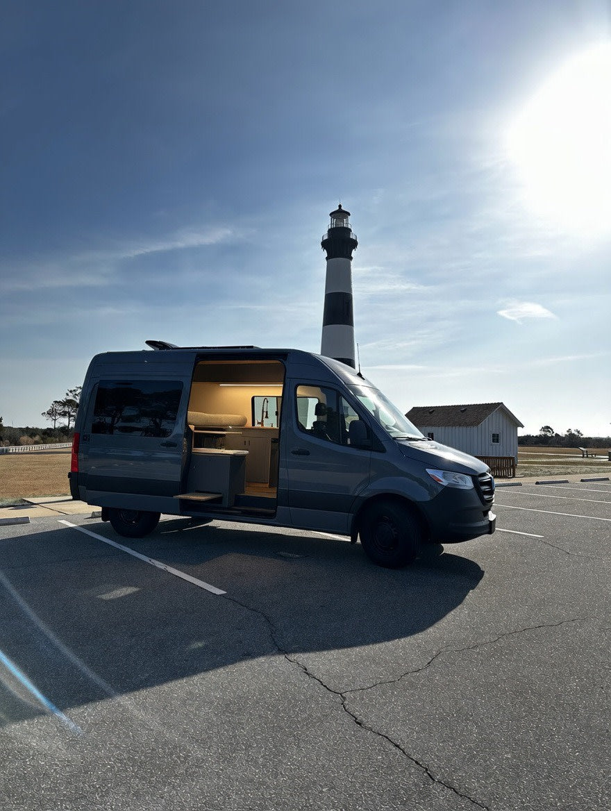 Coastal adventure with an rv rental! A modern gray camper van, door open, interior lit, parked at a lighthouse on a sunny day.