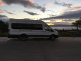 A white RV rental van is parked on asphalt beside calm water at sunset. The sky displays soft pink, orange, and gray clouds.