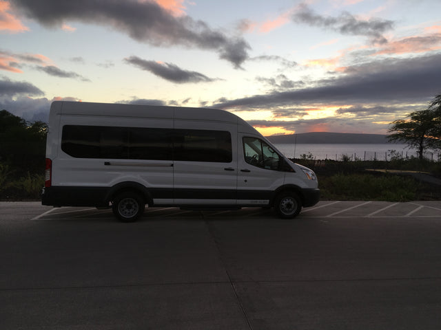 A white RV rental van is parked on asphalt beside calm water at sunset. The sky displays soft pink, orange, and gray clouds.