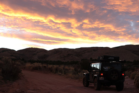A Jeep with a rooftop tent on a dirt trail under a vibrant sunset sky over mountains. Your next RV rental adventure awaits!