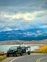 Green Jeep, towing an adventure camper (like an RV rental) with a bike, overlooks a lake and fall mountains.