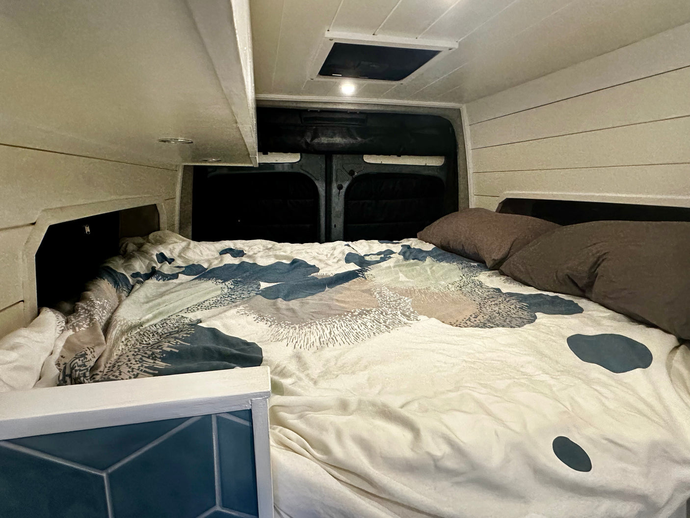 An RV rental's cozy sleeping area features a bed with patterned bedding, built-in storage, and a ceiling vent.