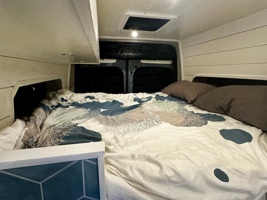 An RV rental's cozy sleeping area features a bed with patterned bedding, built-in storage, and a ceiling vent.