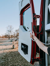 A hand holds a shower sprayer, water streaming from an RV rental van's open door into a field. Ideal for mobile living.