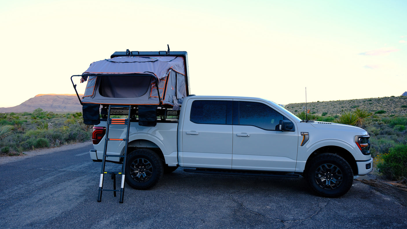 White F-150 truck with an open rooftop tent & ladder parked on a desert road. Perfect for an RV rental or off-road overlanding adventure.