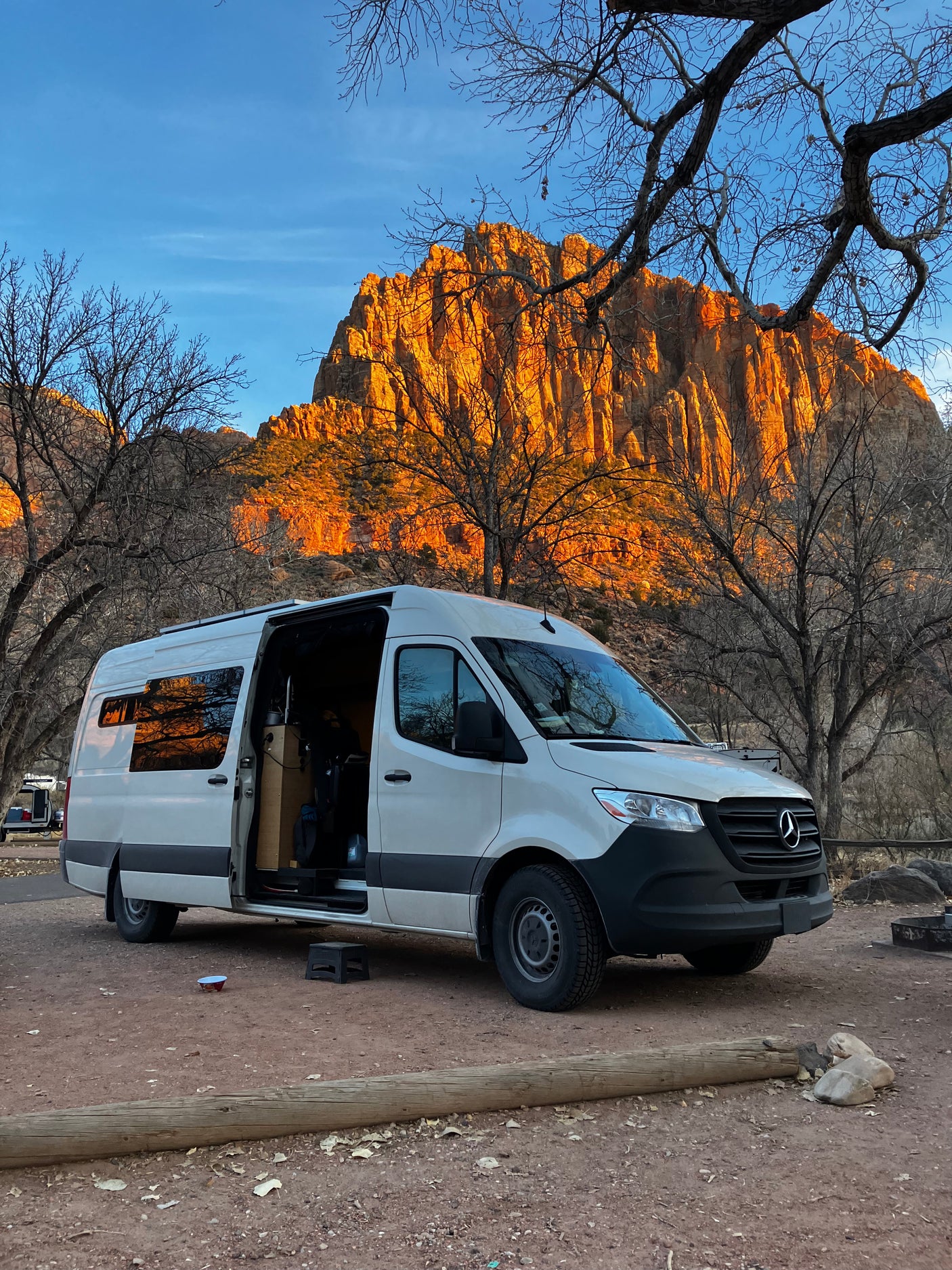 A white camper van, an ideal RV rental, is parked amidst bare trees with a stunning, sunlit golden mountain in the background.