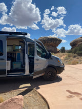 Adventure awaits! This grey rv rental van, side door open to show interior, is parked by massive desert rocks under a blue sky.