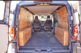 Inside an RV rental van: rear view showing wooden walls, two long benches, & dark carpet. Simple, functional setup.
