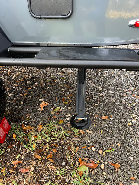 Side of an RV rental showing extended stabilizer jack, step, and gravel ground after rain.